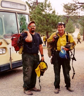 Two firefighters with their gear in front of a bus in a national forest.